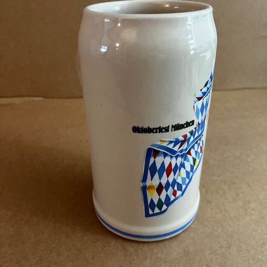 A white ceramic beer stein mug featuring the text 'Oktoberfest München' and a colorful Bavarian flag graphic with blue, red, yellow, and white diamonds, set against a plain brown cardboard background.