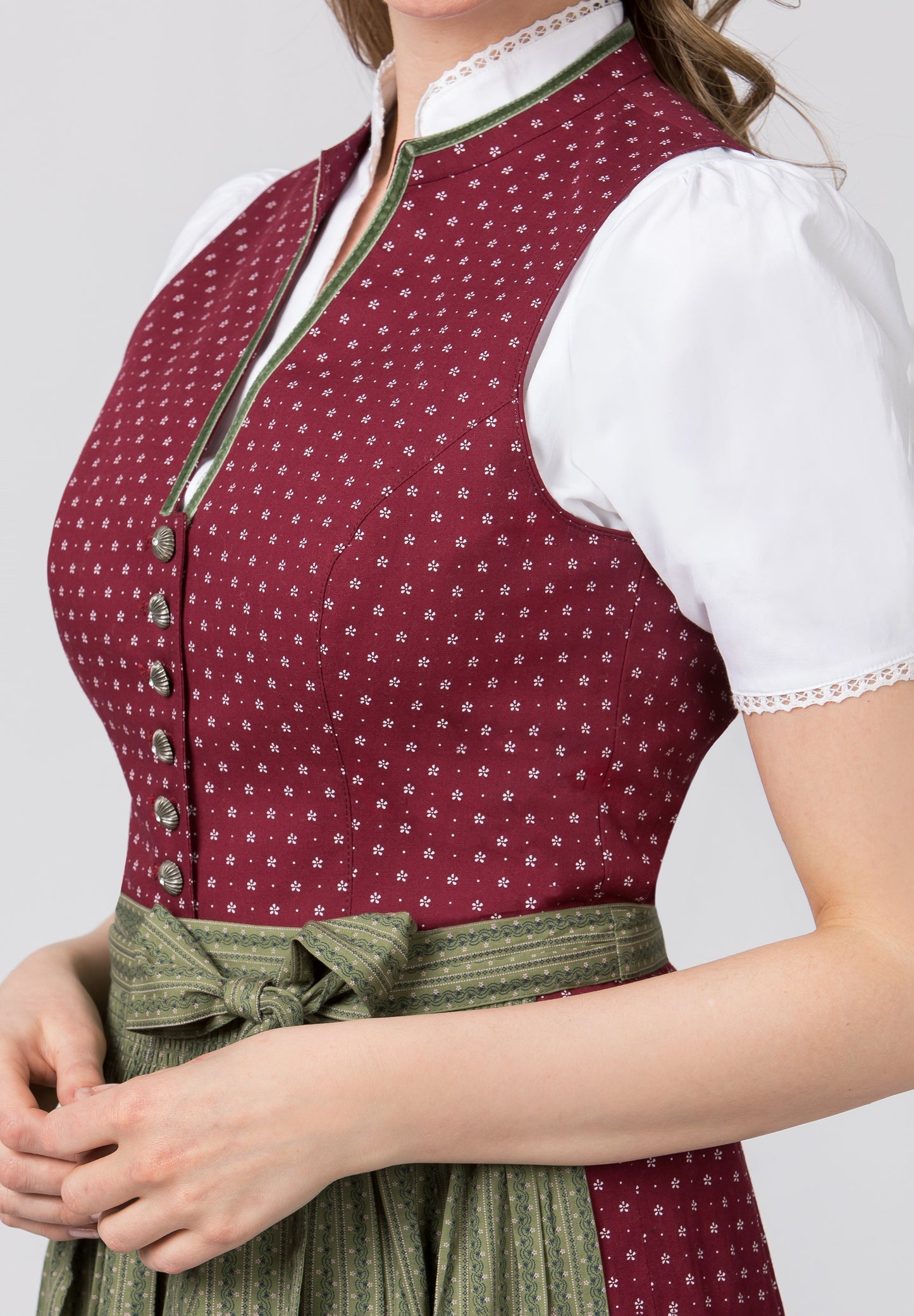A woman wearing a traditional Dirndl dress with a high-necked bodice in bordeaux color, a green apron, and a white blouse with a stand-up collar.