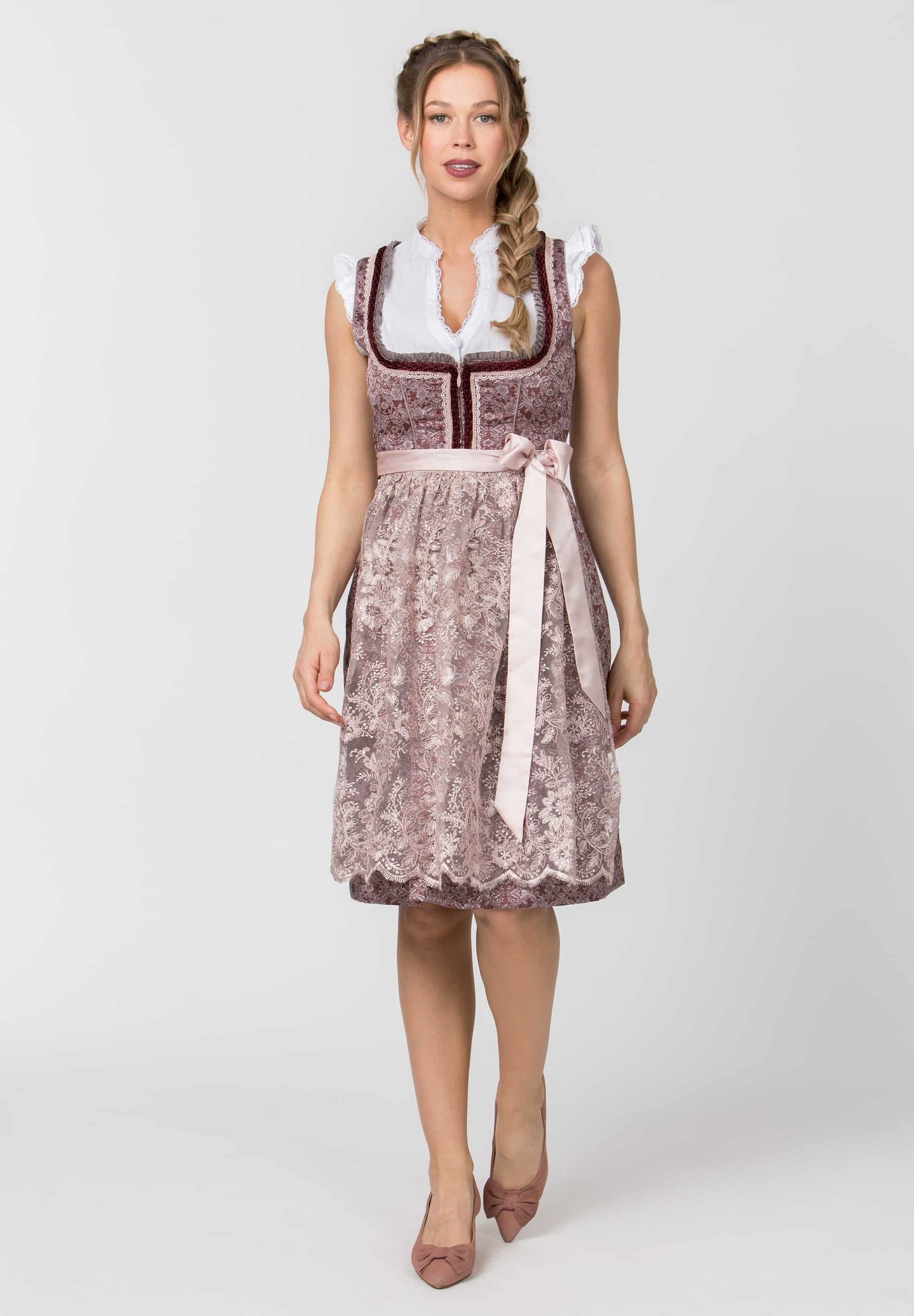 A woman modeling a traditional German dirndl dress with a dusty rose floral pattern, white collar, and a light pink waist sash, photographed against a neutral light gray background.