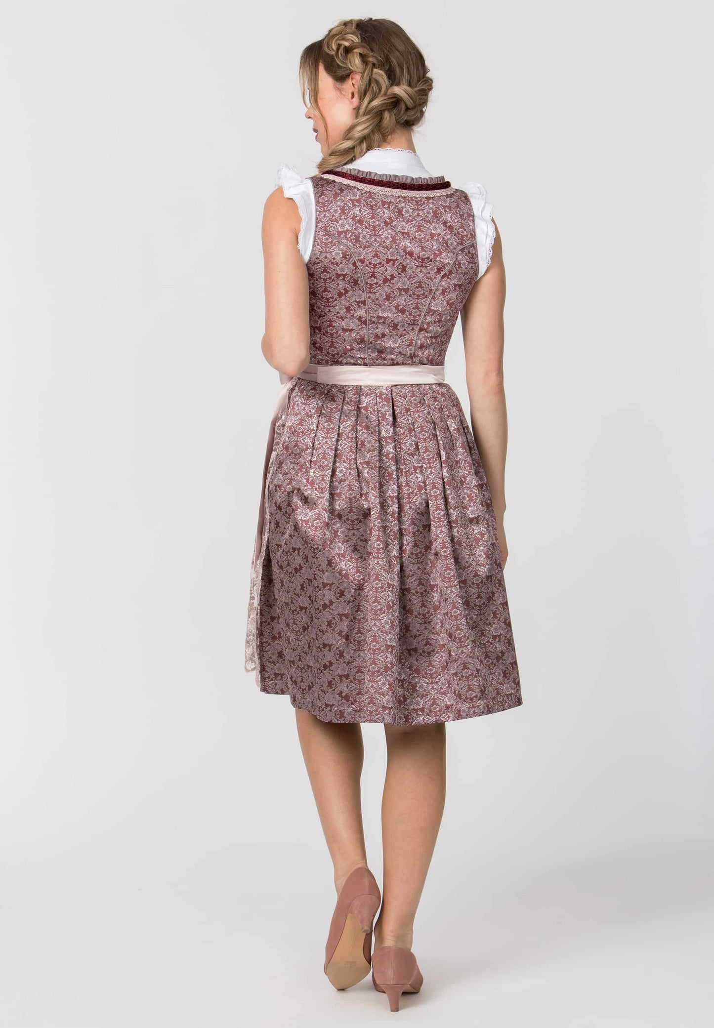 A woman shown from the back wearing a sleeveless midi dress with a floral pattern in dusty pink and purple, featuring white ruffled shoulder details and a white waistband, set against a plain light gray studio background.