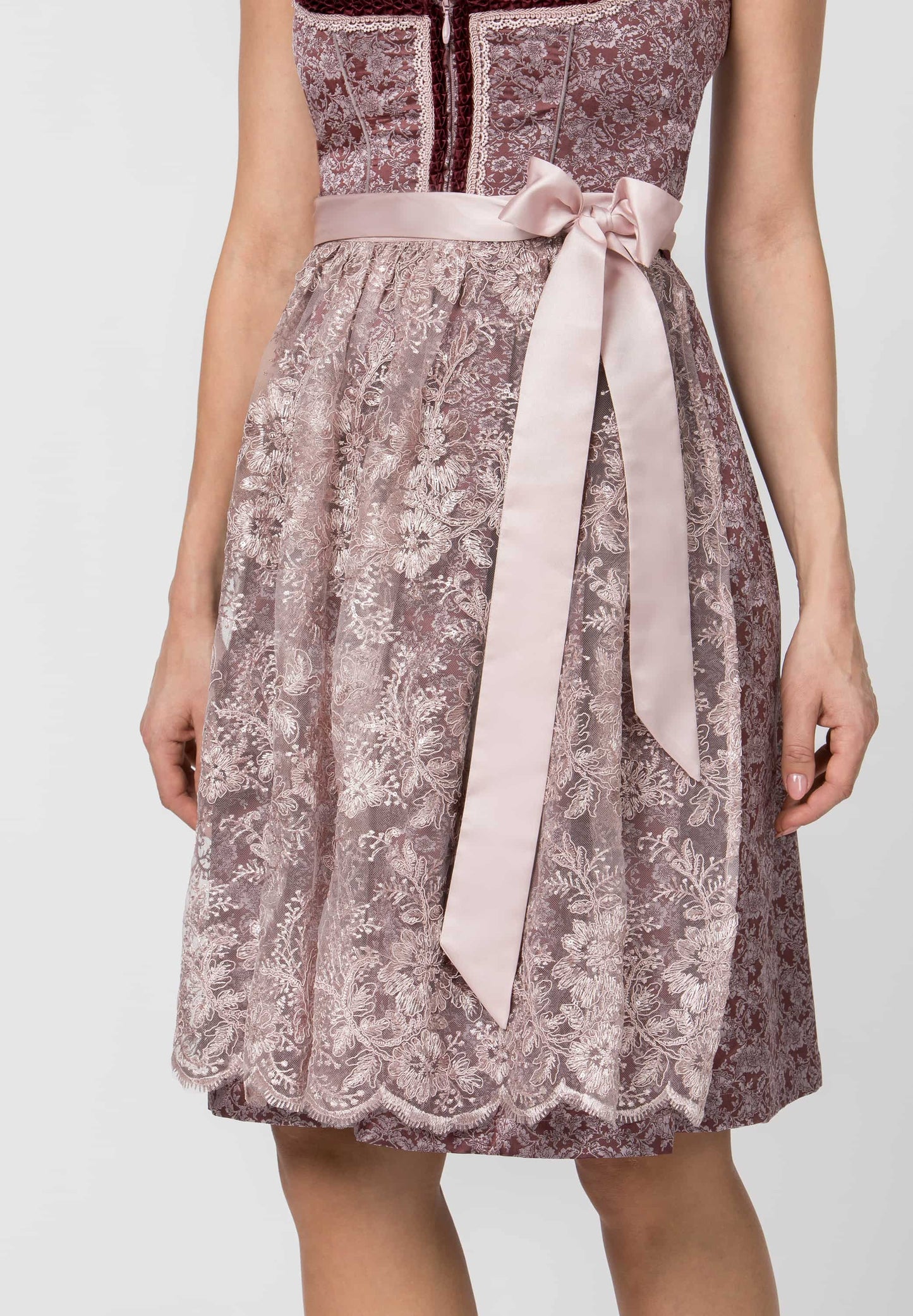 A woman wearing a sleeveless midi dress with a dusty mauve floral lace pattern, a dark burgundy yoke, and a light pink satin waist tie with a long bow.