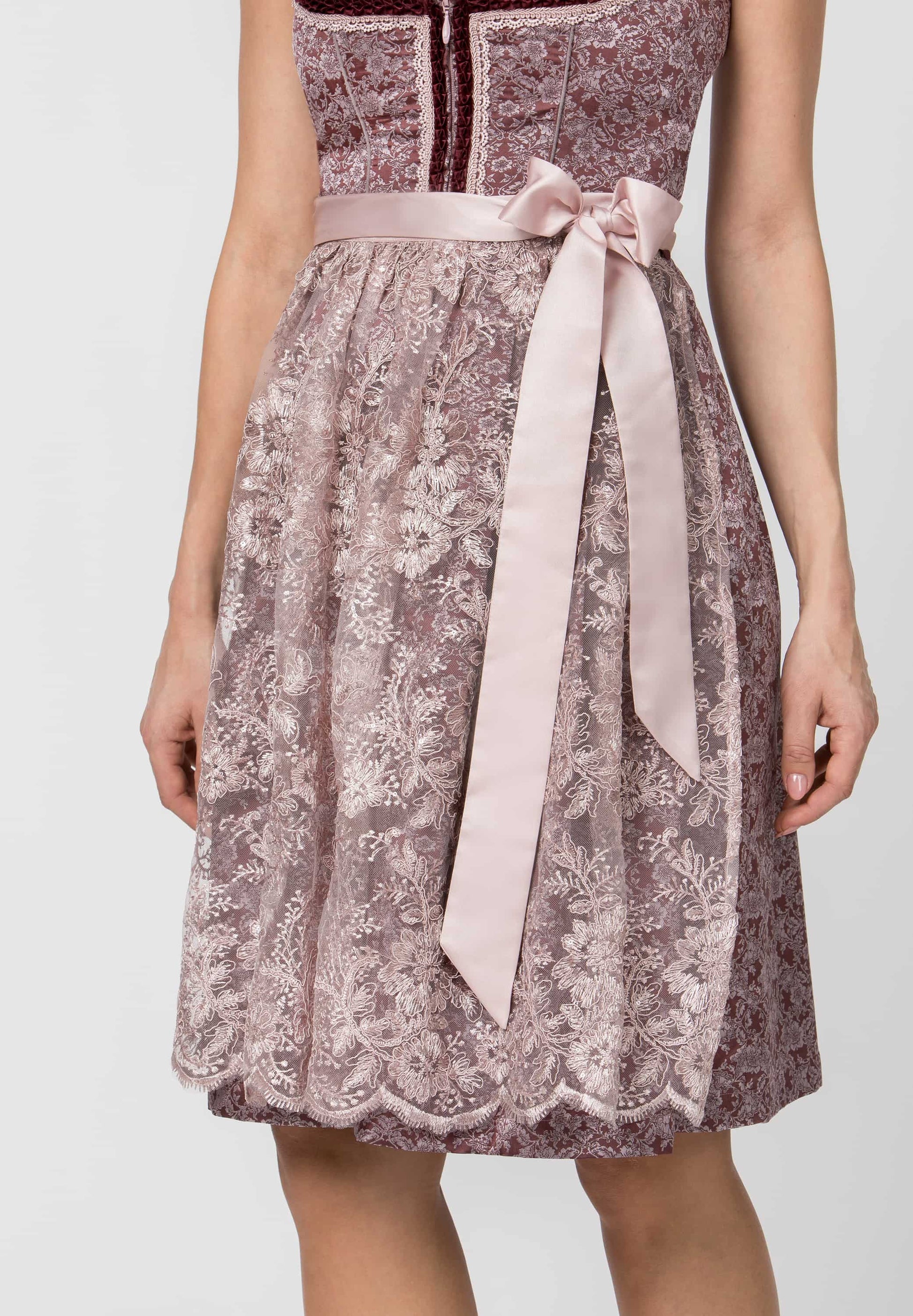 A woman wearing a sleeveless midi dress with a dusty mauve floral lace pattern, a dark burgundy yoke, and a light pink satin waist tie with a long bow.