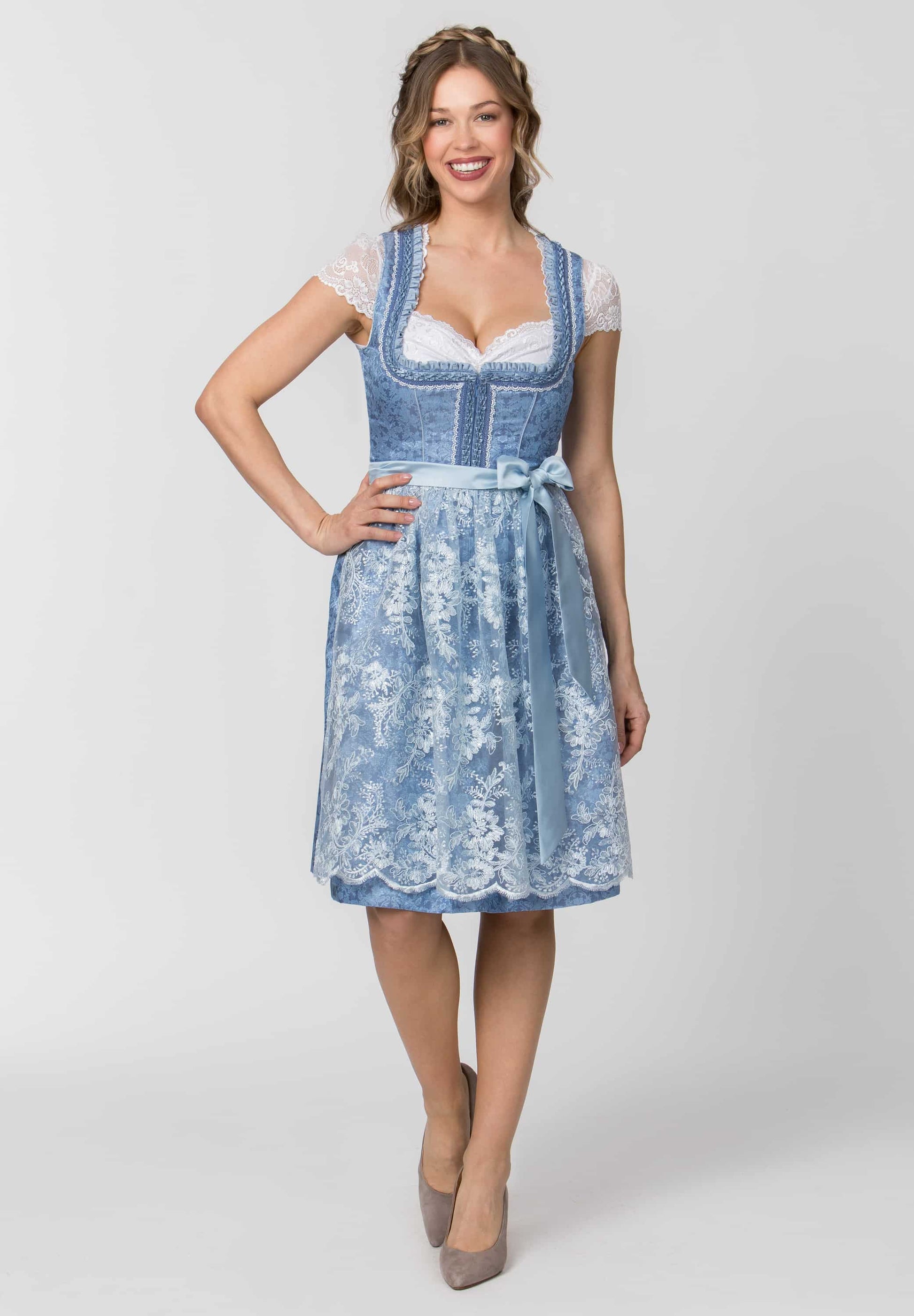 A woman modeling a light blue denim-style dirndl dress with white lace trim, lace sleeves, and a matching waist tie, photographed against a light gray studio background.