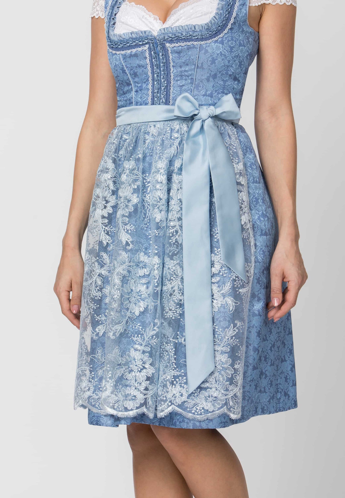 A woman wearing a light blue and white floral-patterned dirndl dress with a matching waist tie, shown from the chest down against a plain light gray background.
