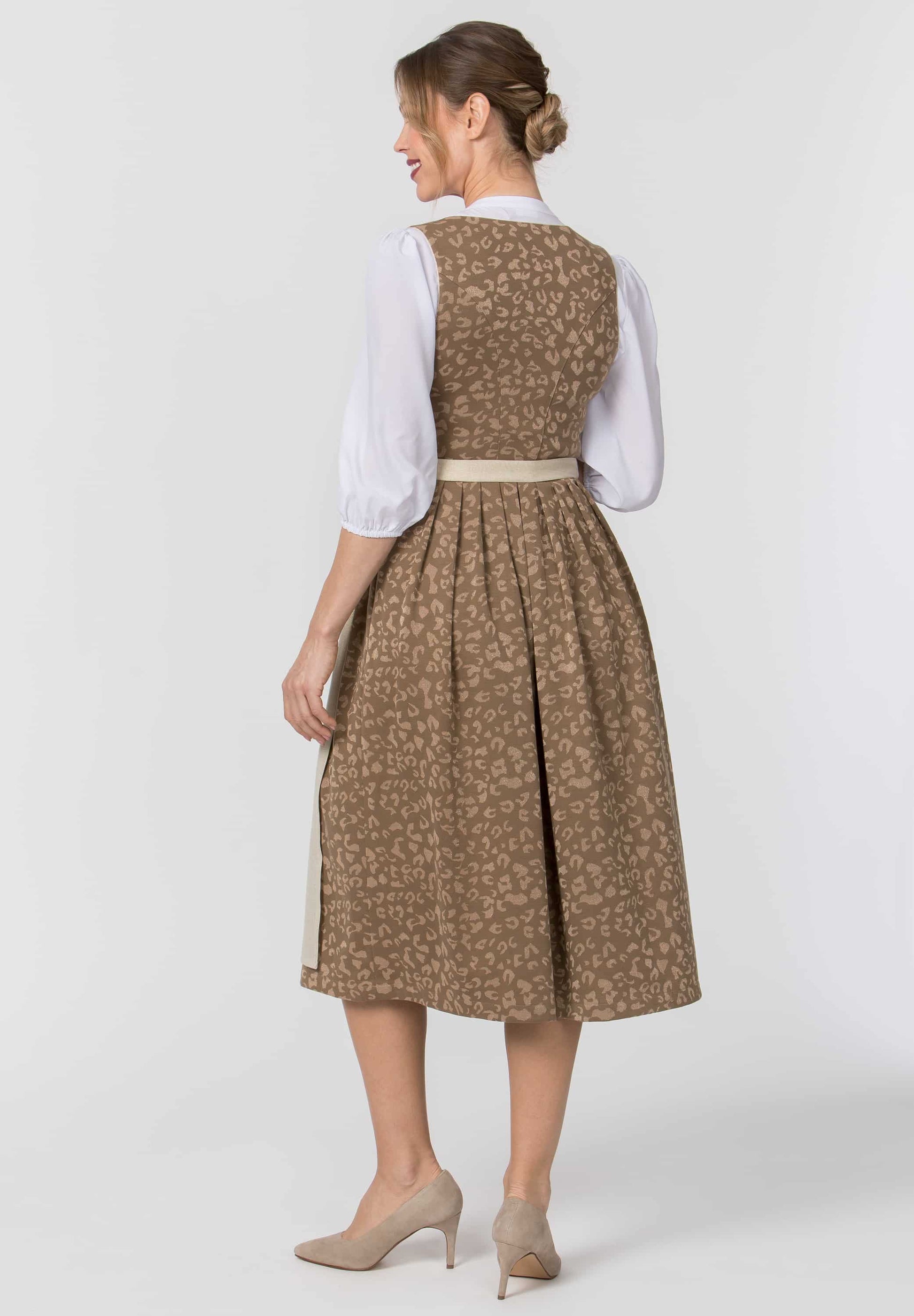 A woman shown from the back wearing a traditional German dirndl dress with a brown and beige floral pattern, a white blouse, and beige heels, photographed against a light gray studio background.