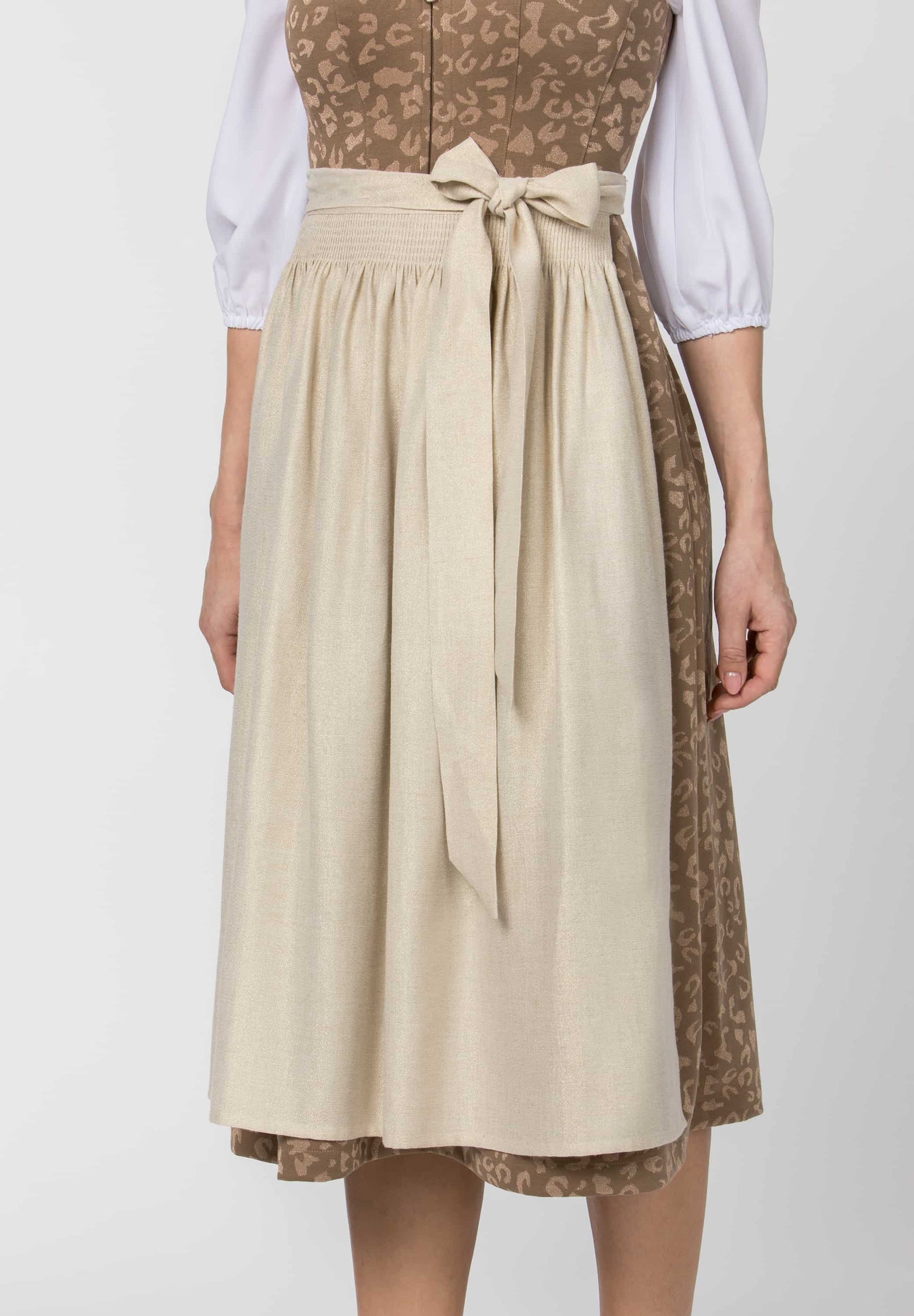 A person wearing a traditional dirndl dress with a beige linen apron skirt tied at the waist, a brown patterned bodice, and white puffed sleeves, shown against a light gray studio background.