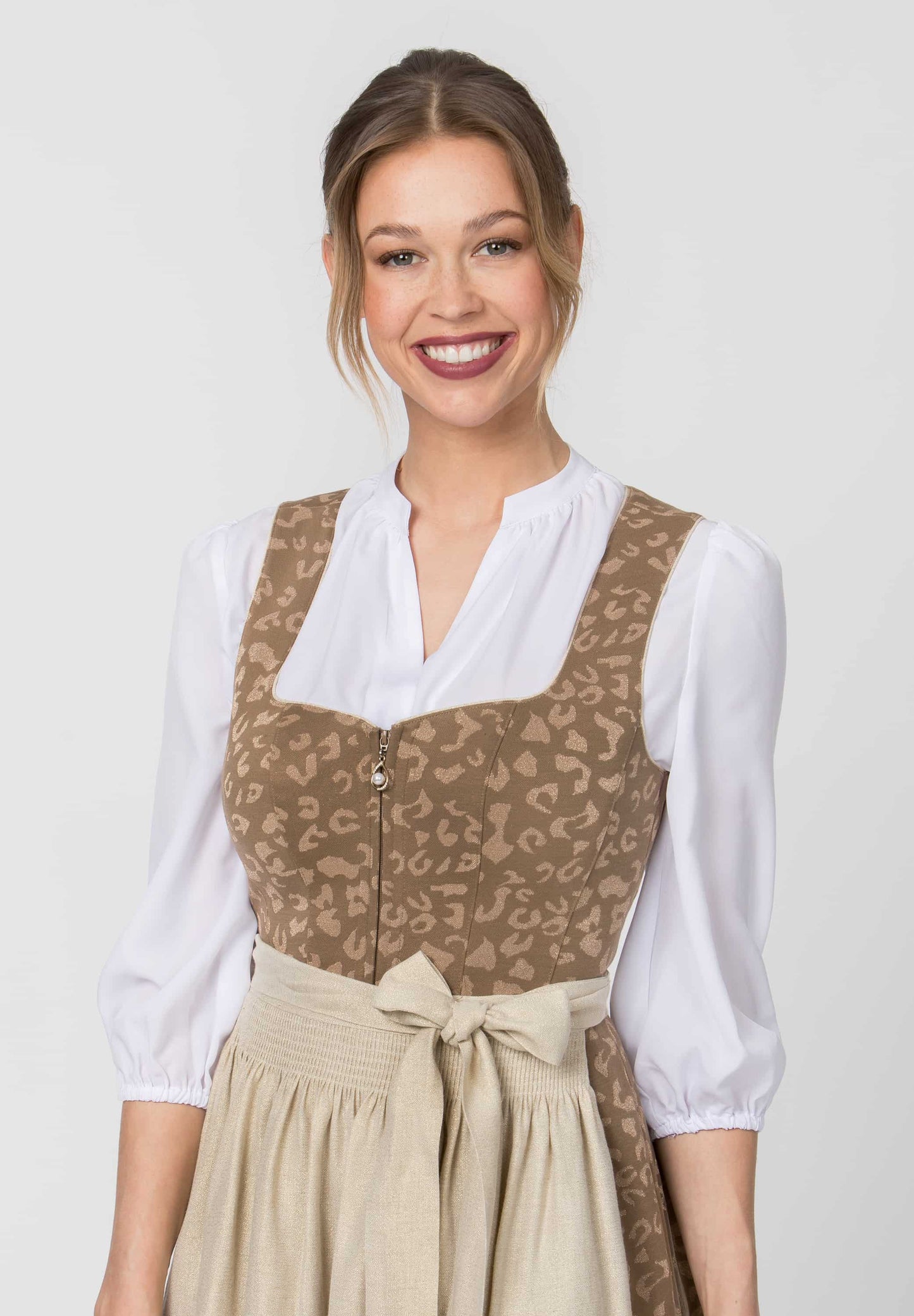 A woman wearing a traditional German dirndl dress with a brown and beige animal print bodice, a white blouse underneath, and a beige skirt with a waist tie, set against a plain light gray background.