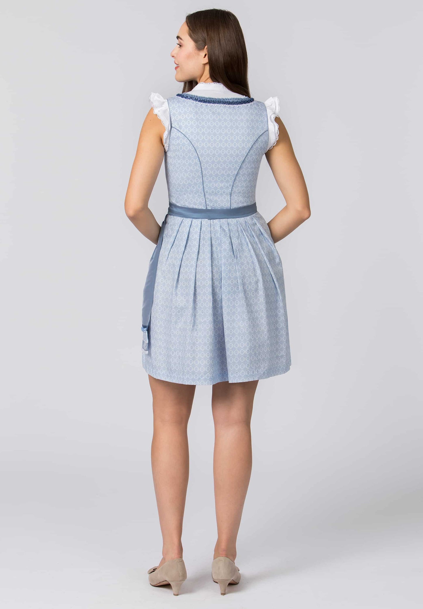 A woman shown from the back wearing a light blue and white patterned dirndl dress with ruffled shoulder details, a blue waistband, and beige heels, photographed against a light gray studio background.