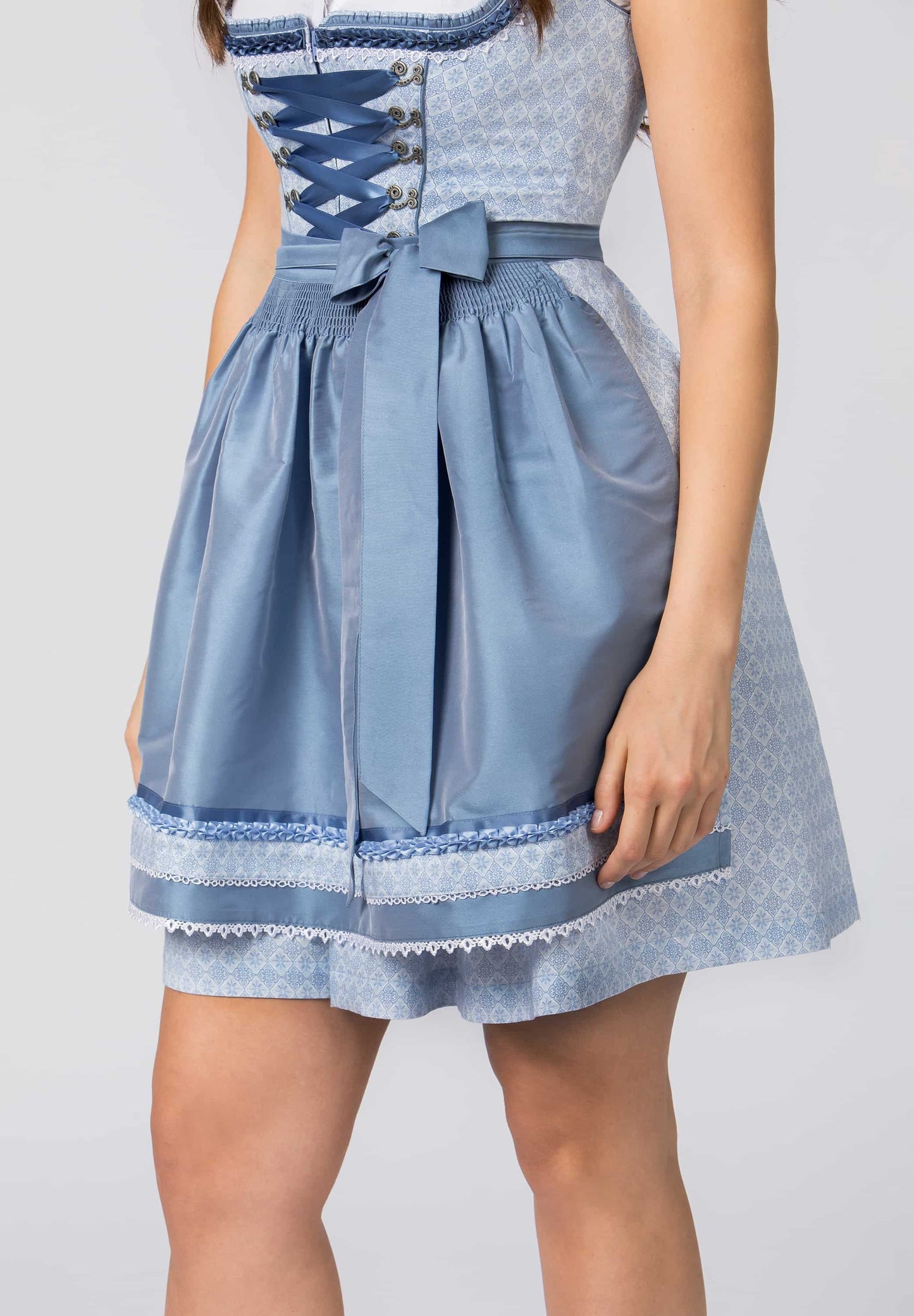 A light blue dirndl dress with a patterned bodice, blue lace-up corset detail, a matching waist tie with bow, and a flared skirt, shown on a model against a light gray background.