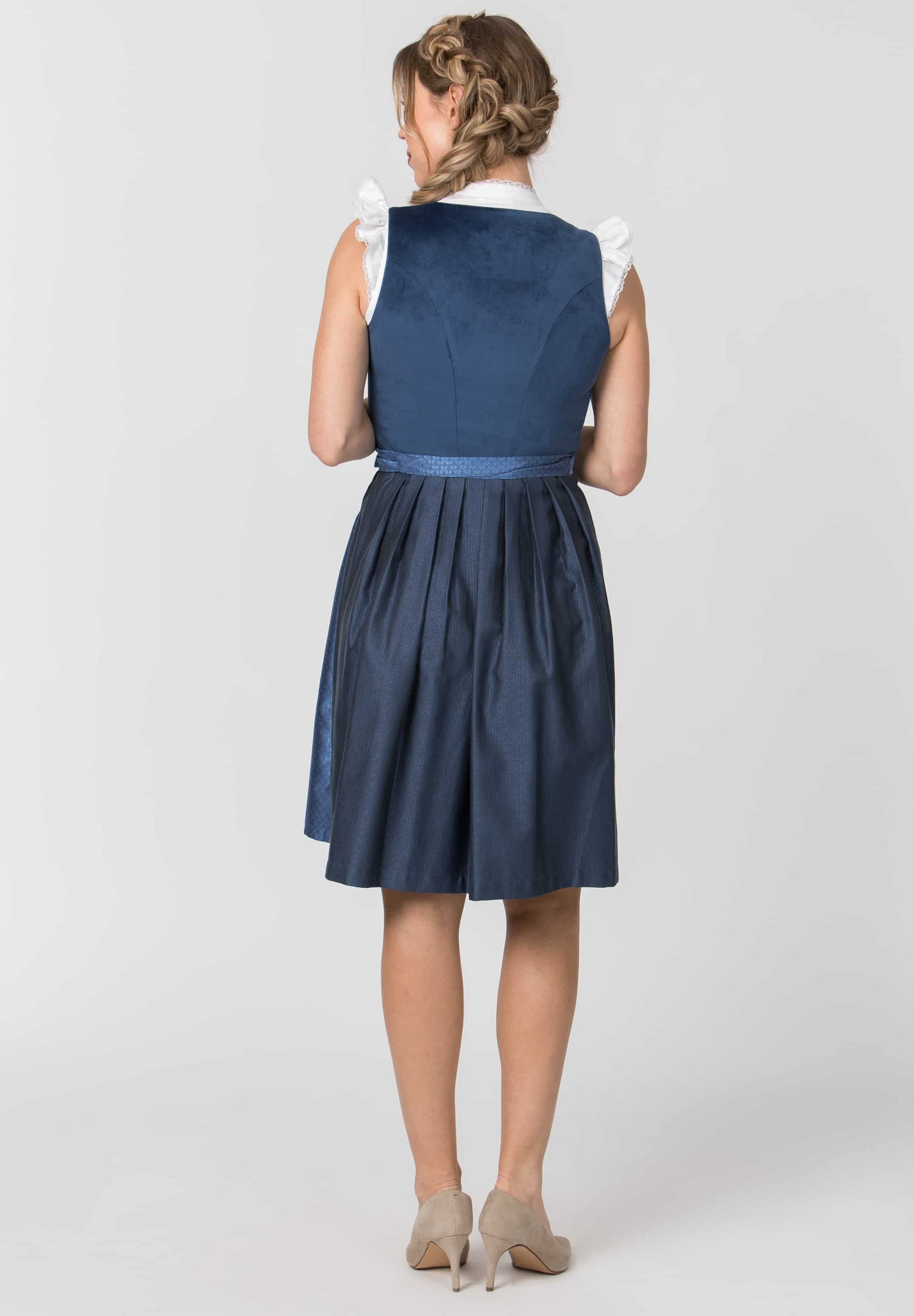 A woman shown from the back wearing a sleeveless navy blue dress with a white ruffled collar and a matching navy waistband, paired with beige heels, against a light gray studio background.