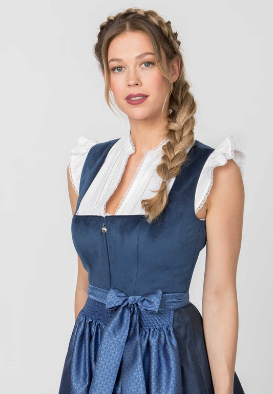 A woman wearing a traditional German dirndl dress with a navy blue body, white pleated collar, and ruffled sleeves, photographed against a light gray studio background.