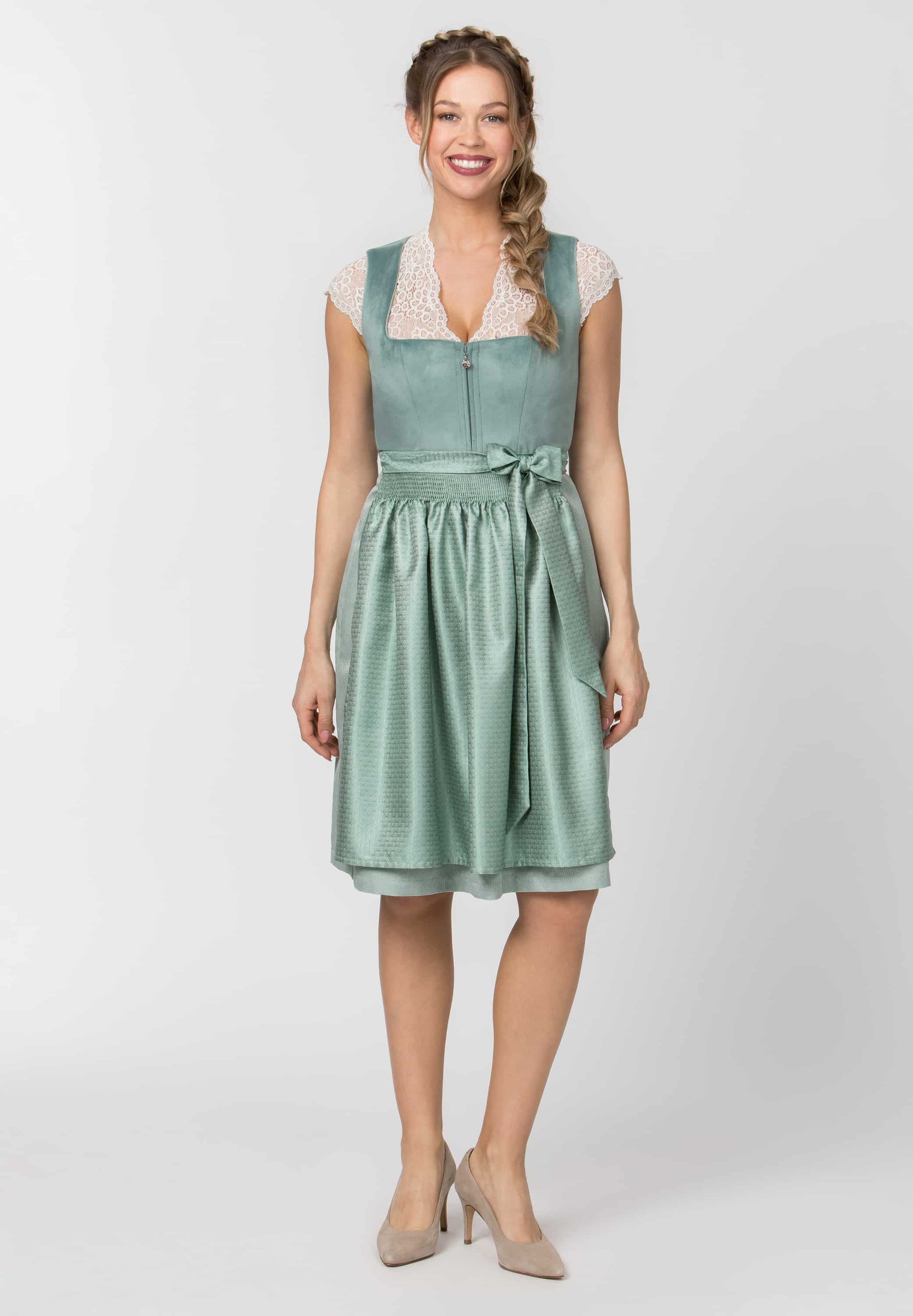 A woman modeling a sage green sleeveless midi dress with a lace V-neckline, tie waist, and gathered skirt, paired with beige heels against a light gray studio background.