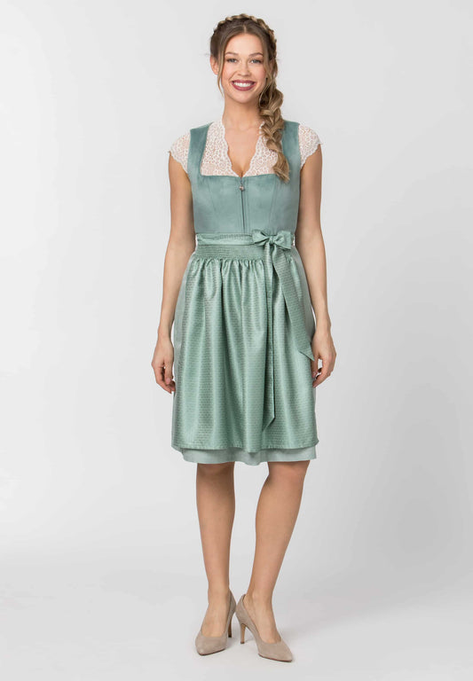 A woman modeling a sage green sleeveless midi dress with a lace V-neckline, tie waist, and gathered skirt, paired with beige heels against a light gray studio background.
