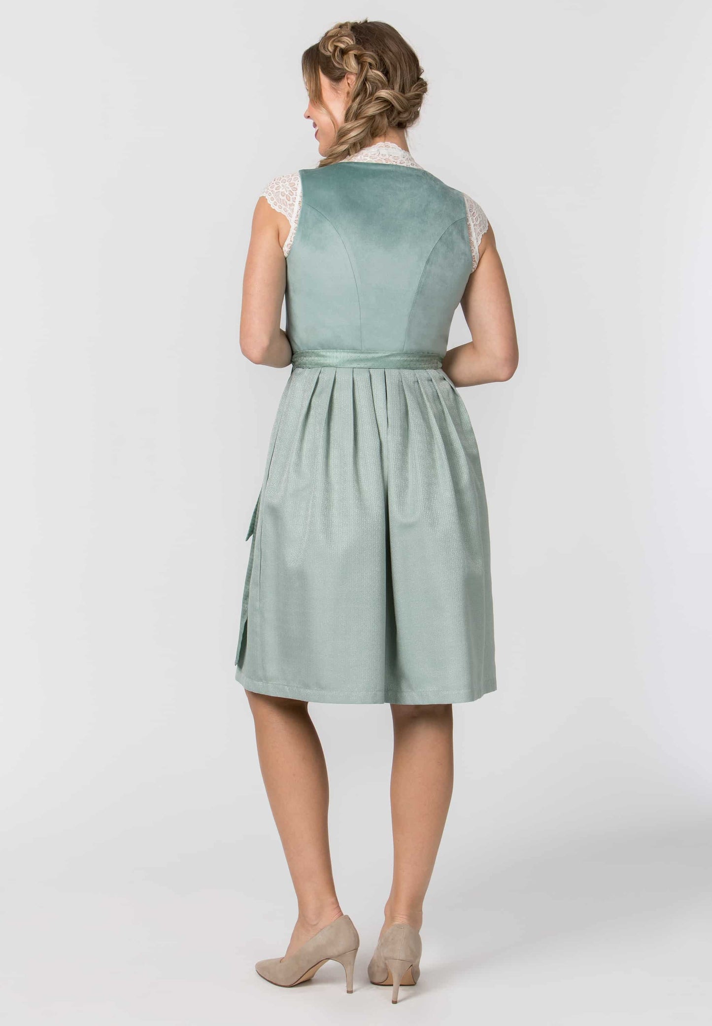 A woman shown from the back wearing a sleeveless sage green dress with a gathered waist and pleated skirt, paired with beige heels, against a light gray studio background.