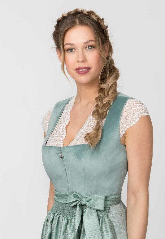 A woman modeling a sage green dirndl dress with white lace trim and a matching waist tie, set against a plain light gray background.