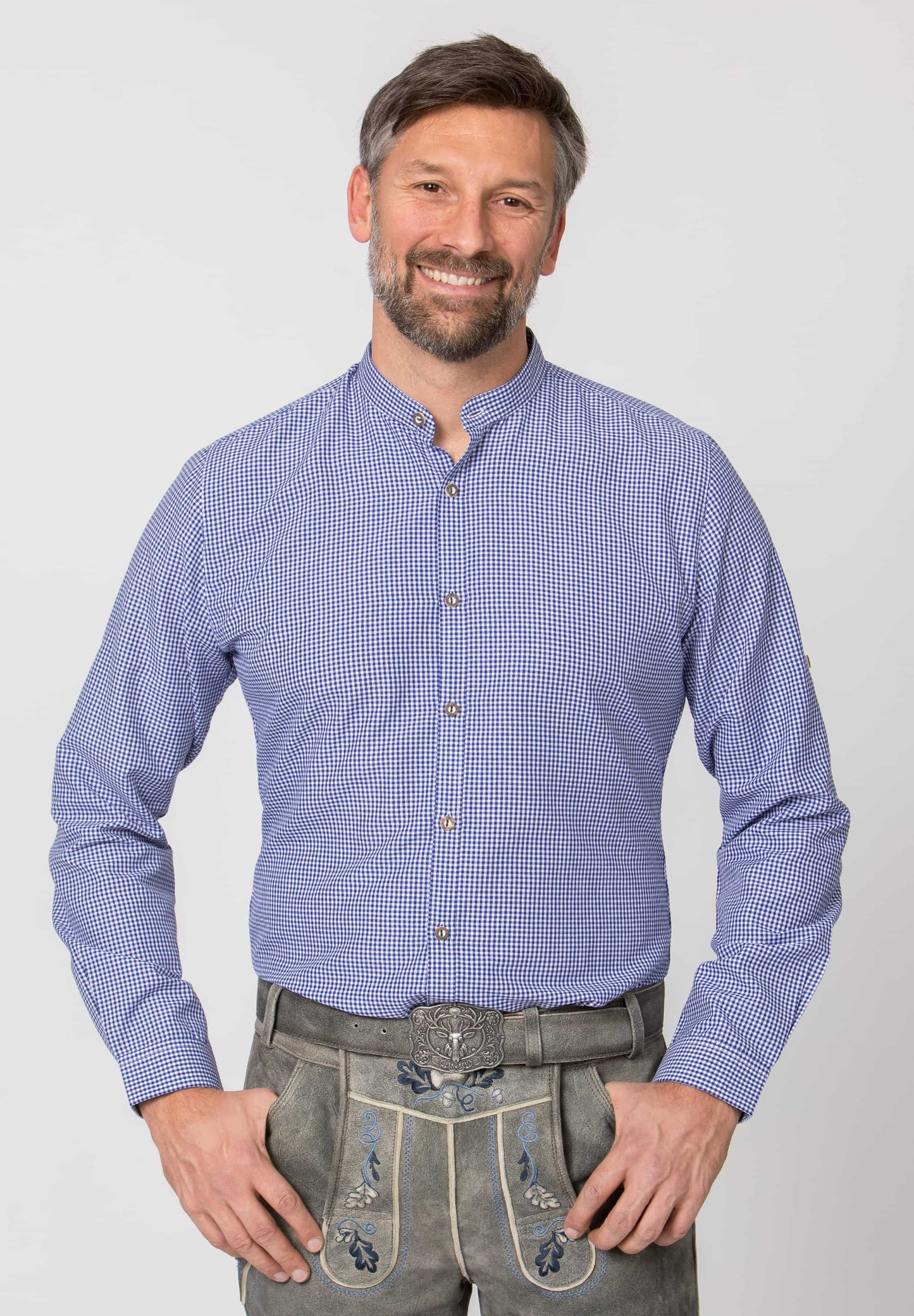 Man wearing a blue checkered shirt and traditional gray pants with floral embroidery on a light gray background