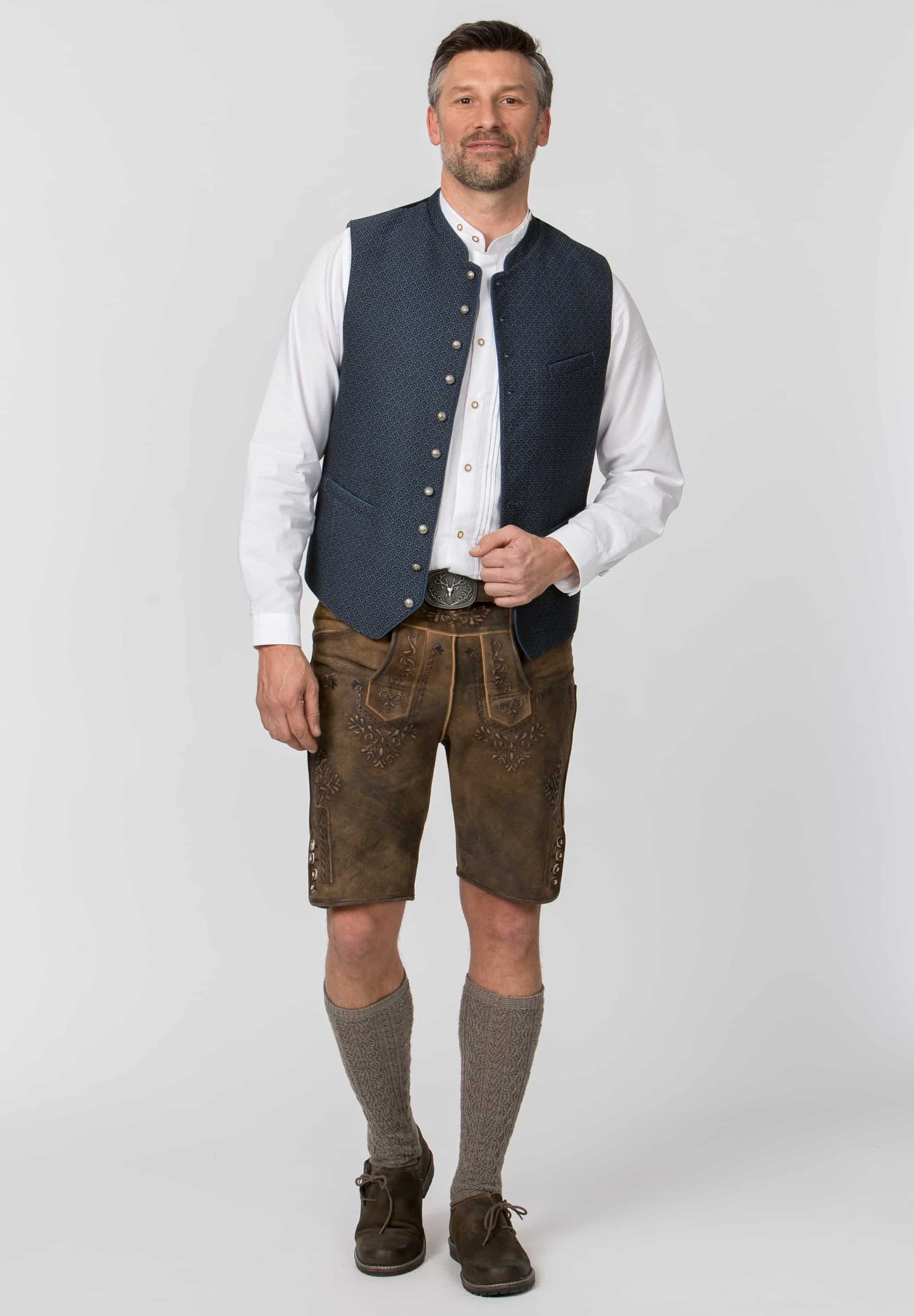 A man modeling traditional Bavarian-style clothing, featuring a dark blue quilted waistcoat over a white long-sleeve shirt, brown leather Lederhosen shorts, gray knee-high socks, and brown leather shoes, against a light gray studio background.