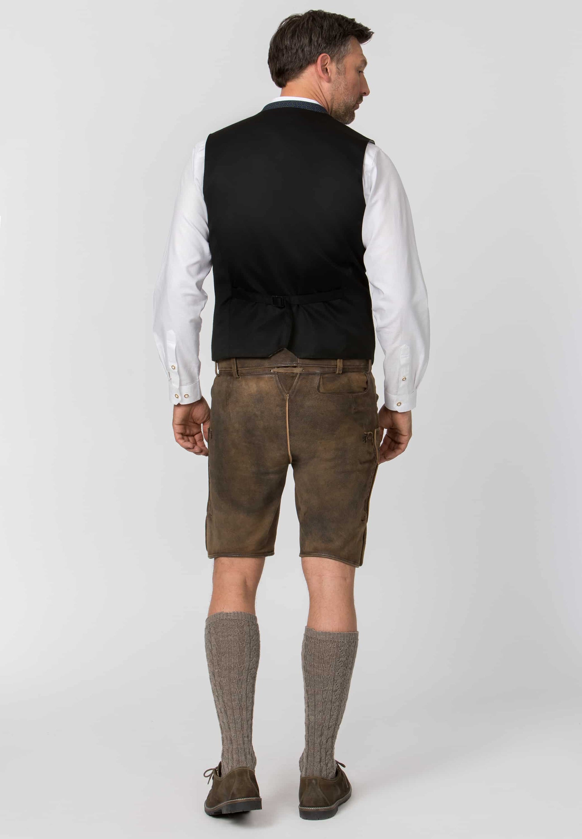 A man shown from the back wearing traditional Bavarian-style clothing: a black waistcoat over a white dress shirt, brown Lederhosen shorts, brown knee-high socks, and brown shoes, against a light gray studio background.