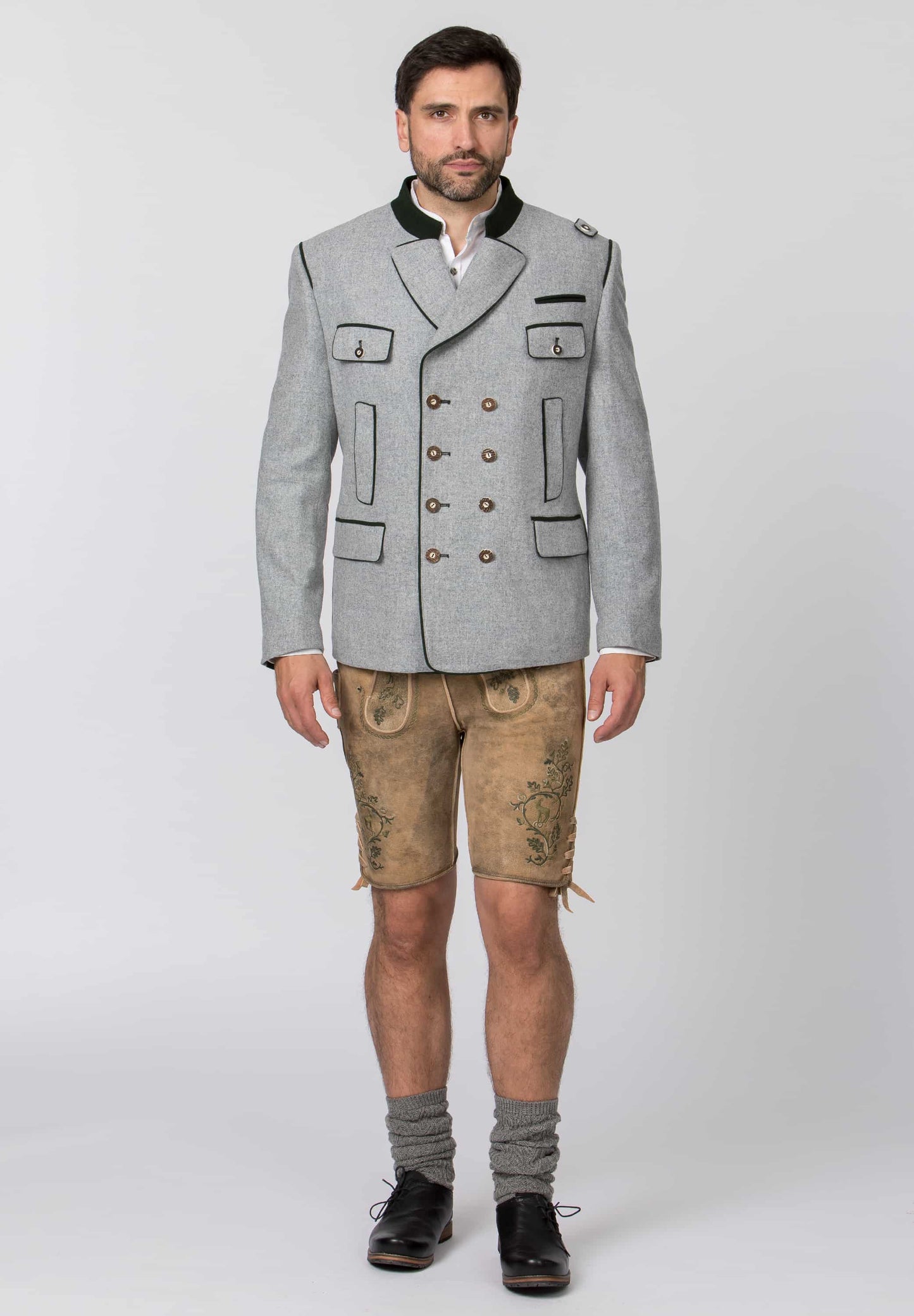 A man modeling a traditional German-style outfit consisting of a light gray double-breasted jacket with black trim, tan embroidered shorts, gray socks, and black shoes, photographed against a neutral light gray background.