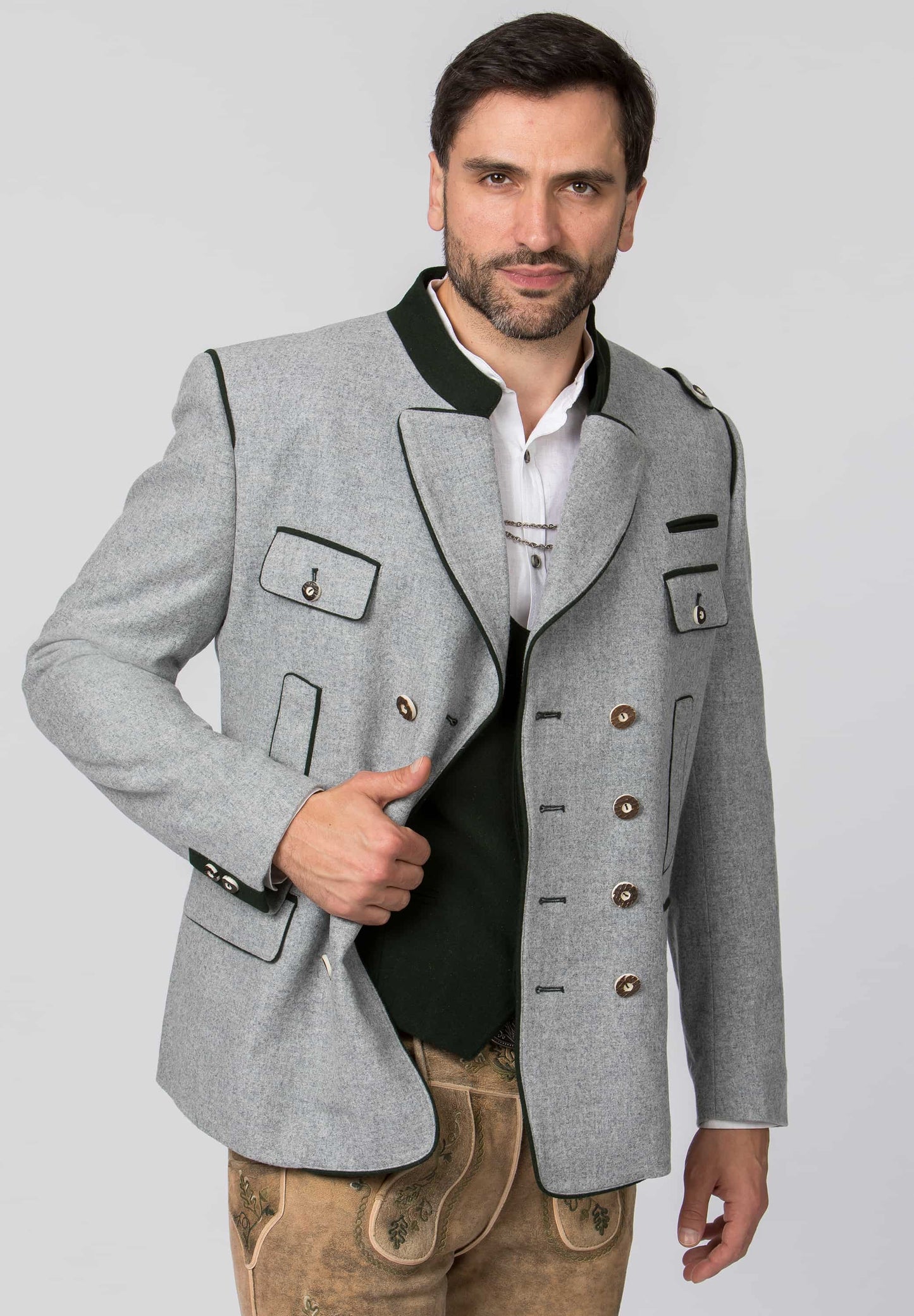 A man modeling a light gray traditional Bavarian-style jacket with black trim, brown buttons, and a white shirt underneath, paired with tan leather pants, against a solid light gray background.