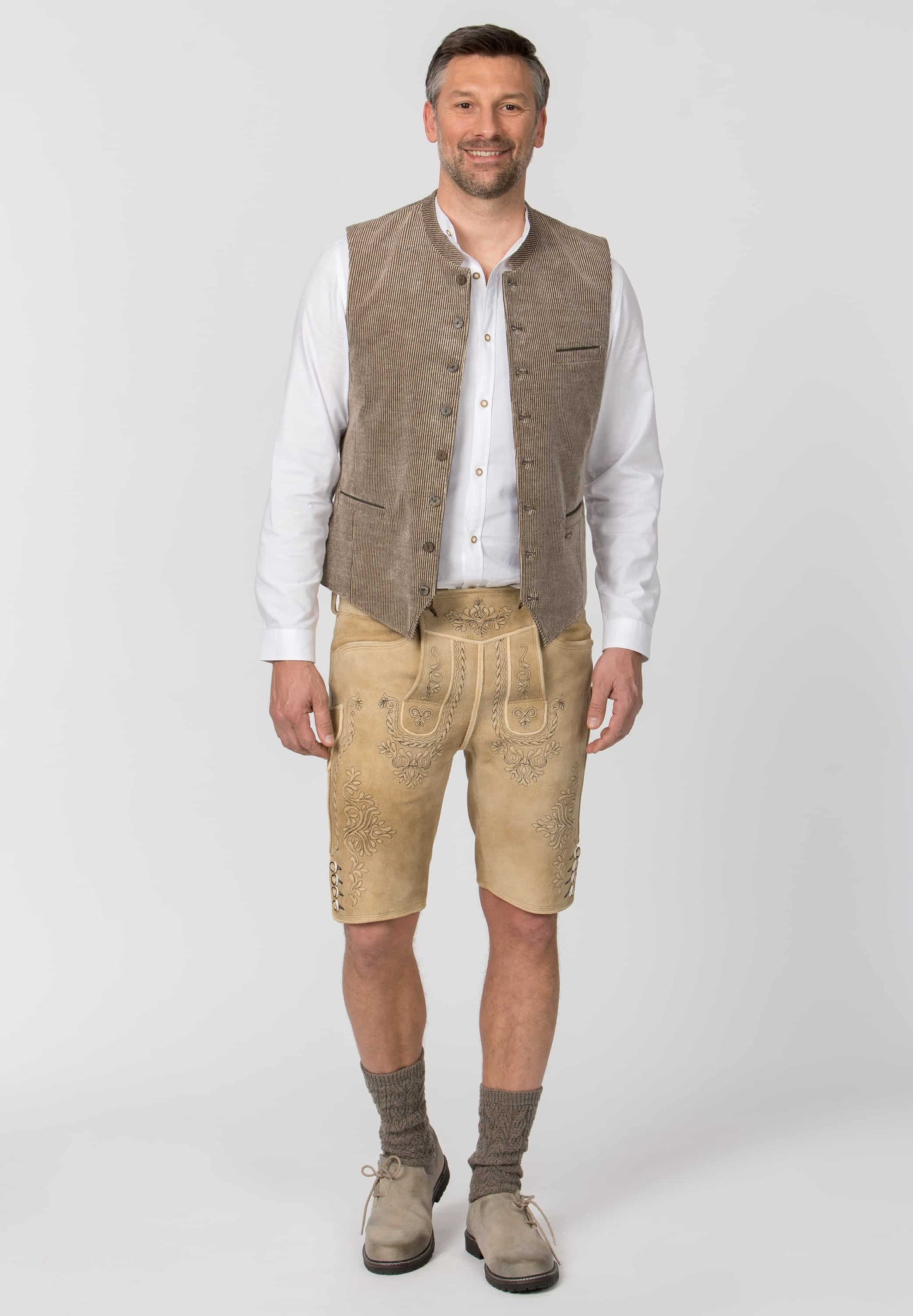 A man modeling traditional Bavarian-style clothing, featuring a brown suede vest, beige embroidered leather shorts, a white button-up shirt, and brown suede boots, shot against a light gray studio background.