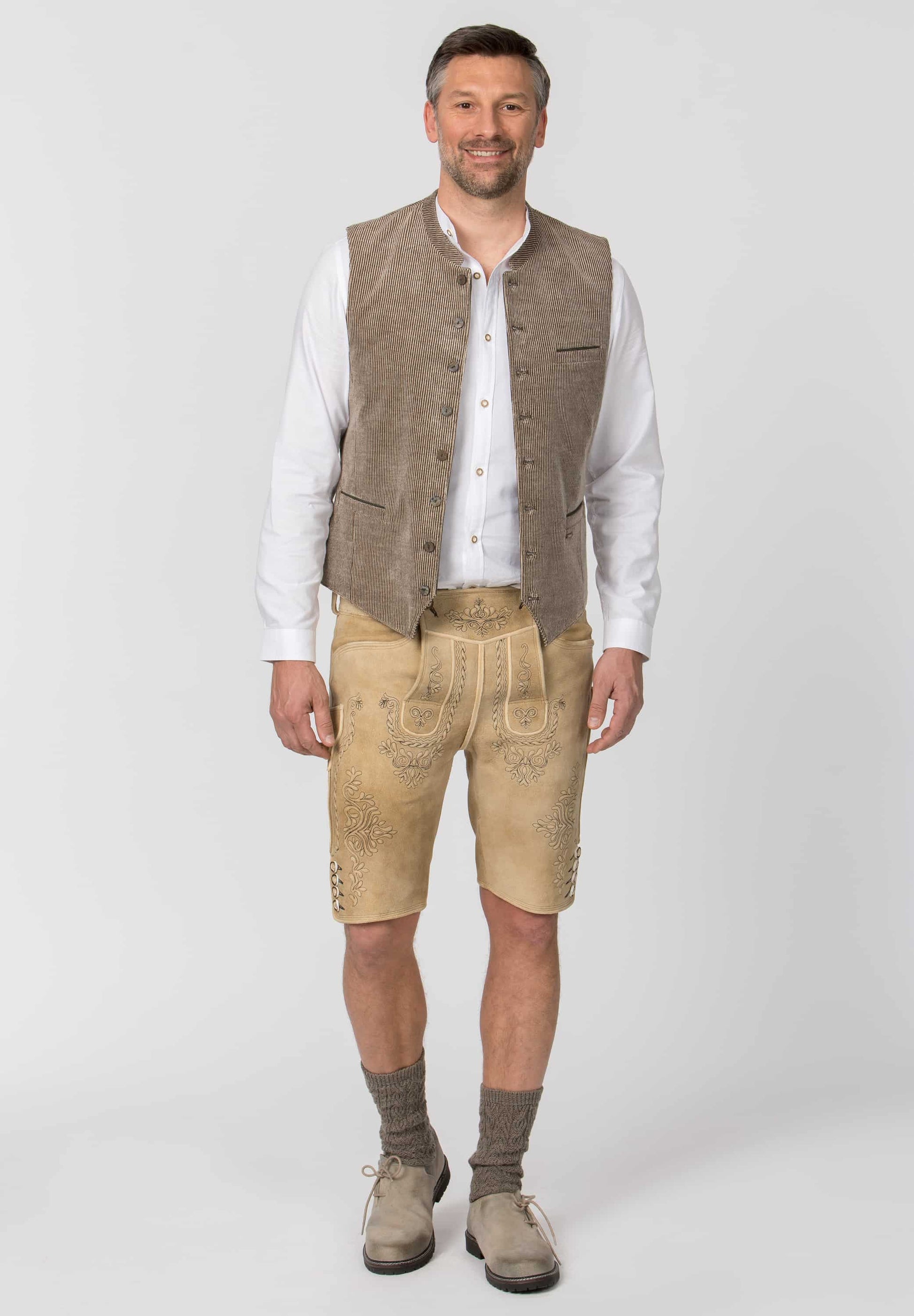 A man modeling traditional Bavarian-style clothing, featuring a brown suede vest, beige embroidered leather shorts, a white button-up shirt, and brown suede boots, shot against a light gray studio background.