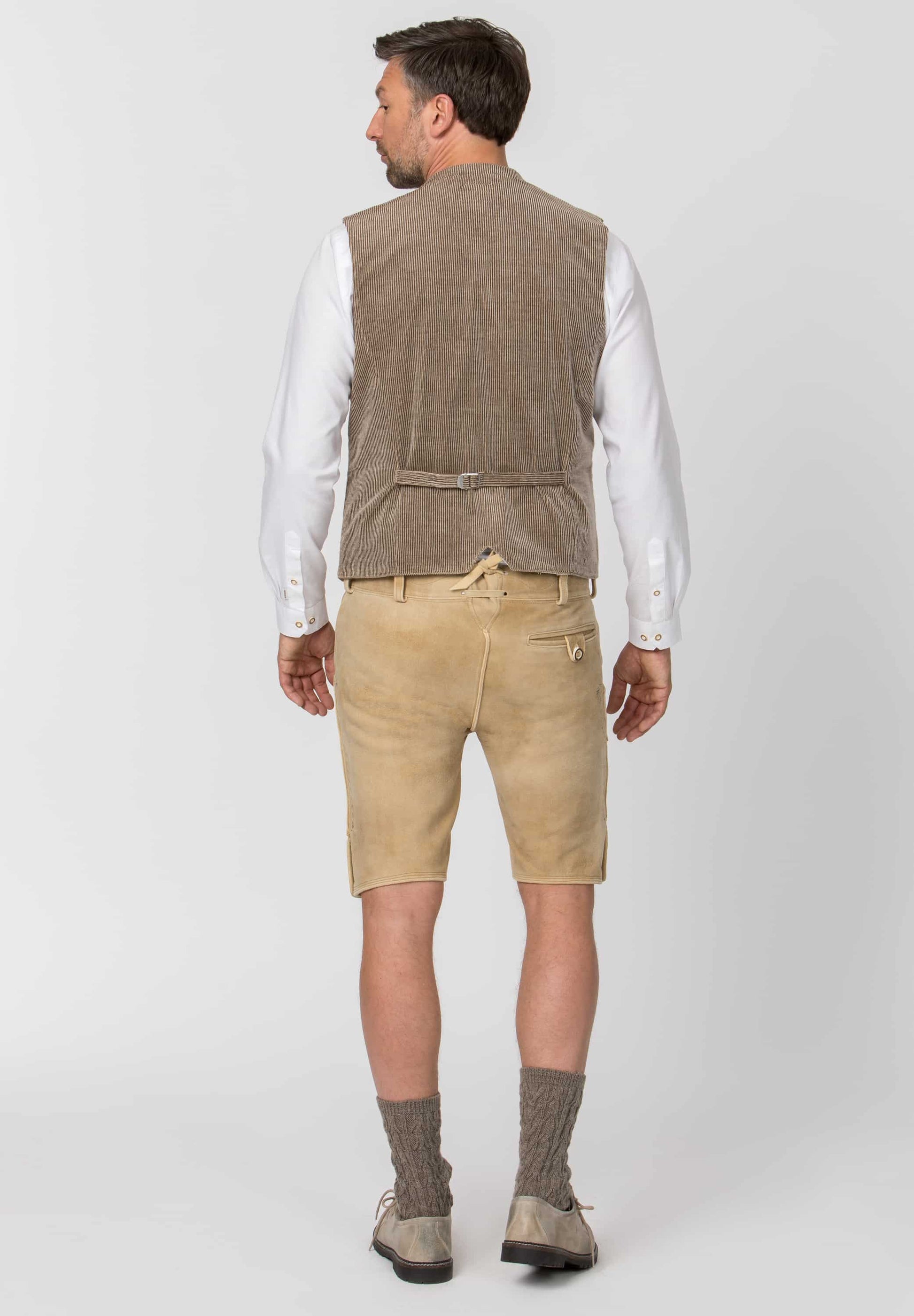 A man shown from the back wearing a brown corduroy vest over a white long-sleeve shirt, paired with beige shorts, brown socks, and brown shoes, against a light gray studio background.