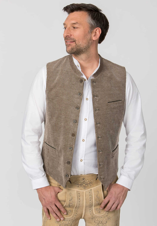 A man wearing a brown textured waistcoat over a white long-sleeve shirt and beige embroidered trousers, photographed against a light gray studio background.