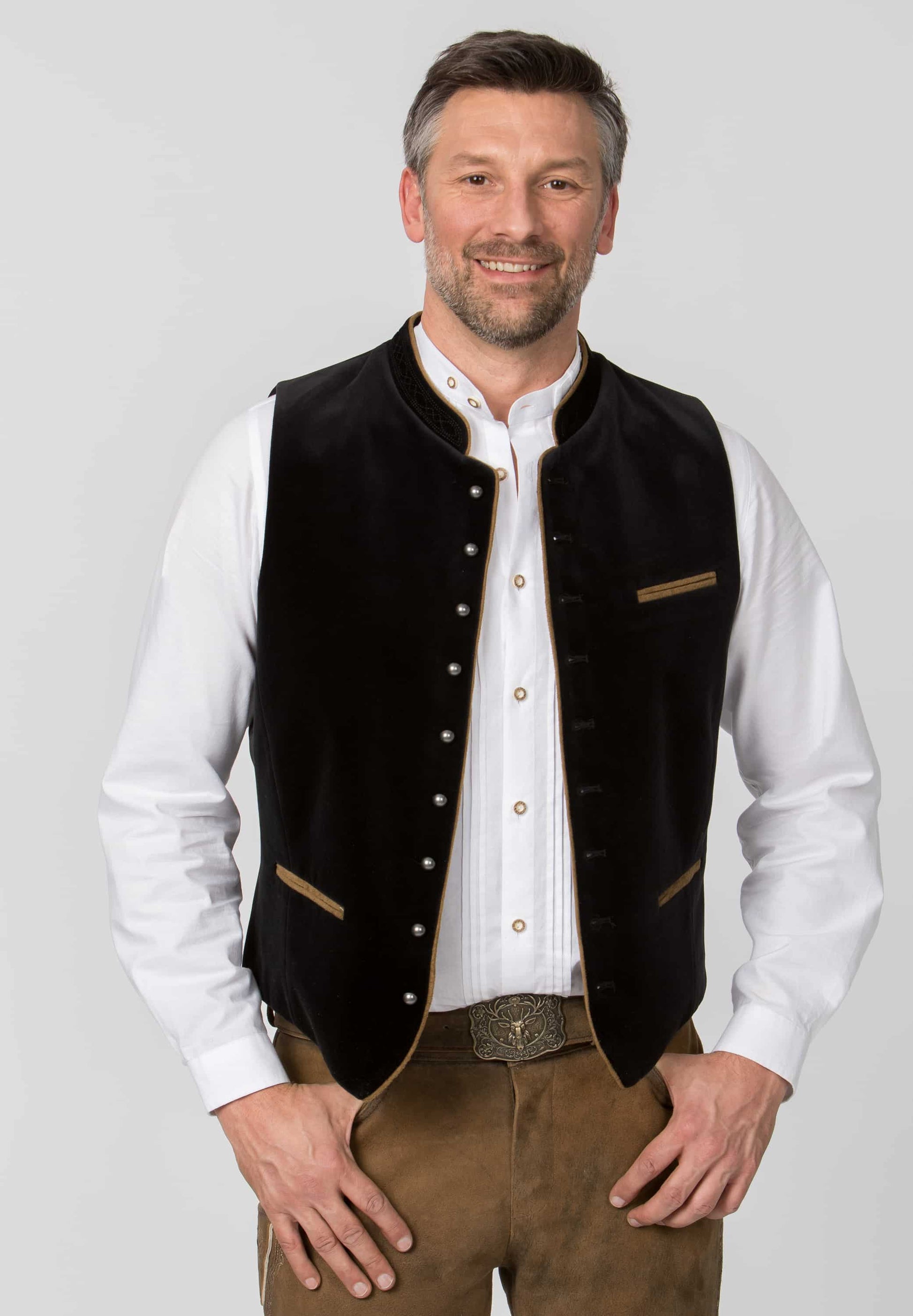 Man wearing a black vest over a white shirt with brown pants on a gray background