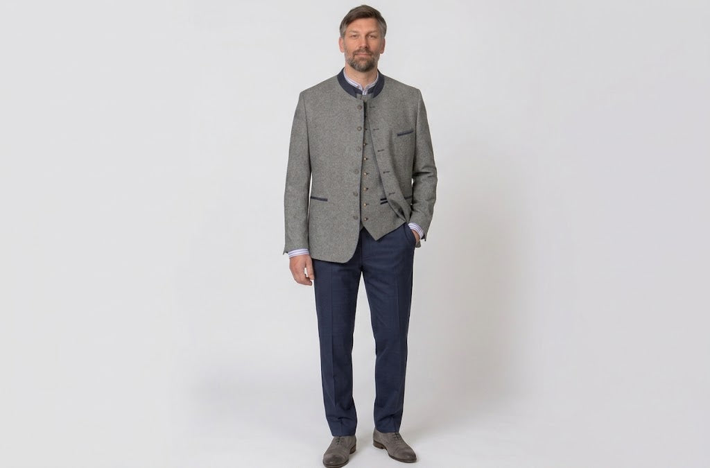 A man modeling a traditional German outdoor jacket (Lederhosenblazer) in a gray herringbone pattern, worn over a matching gray waistcoat, navy trousers, and brown shoes, against a plain light gray background.